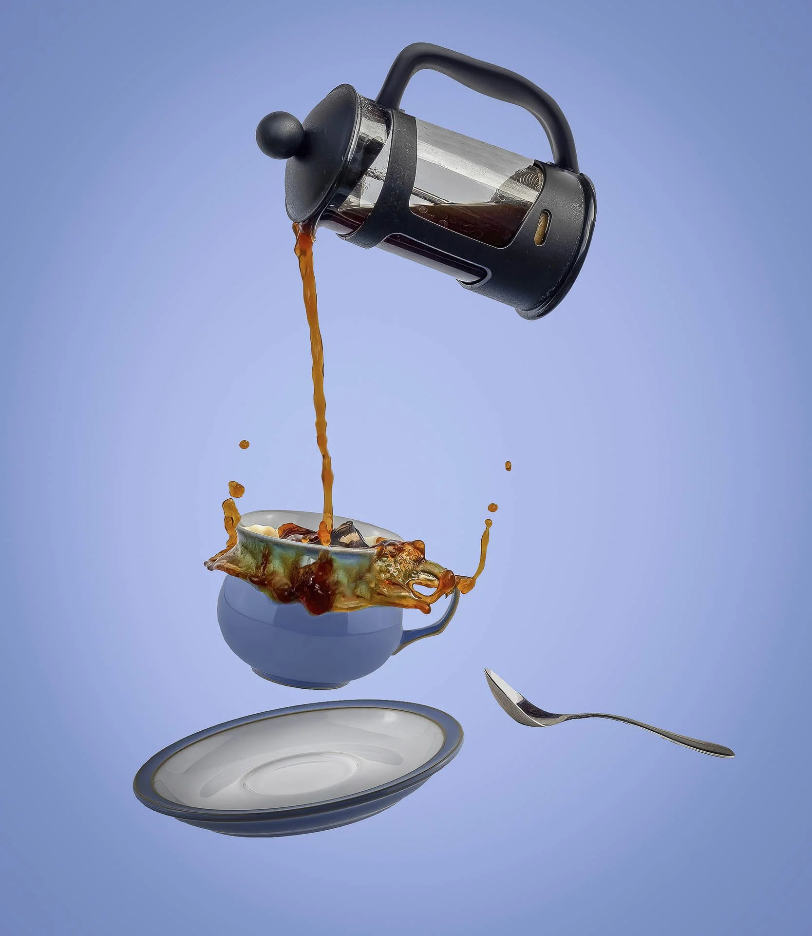 A French press pouring coffee into a blue teacup, causing a splash. The cup is floating above a saucer and spoon against a blue background.