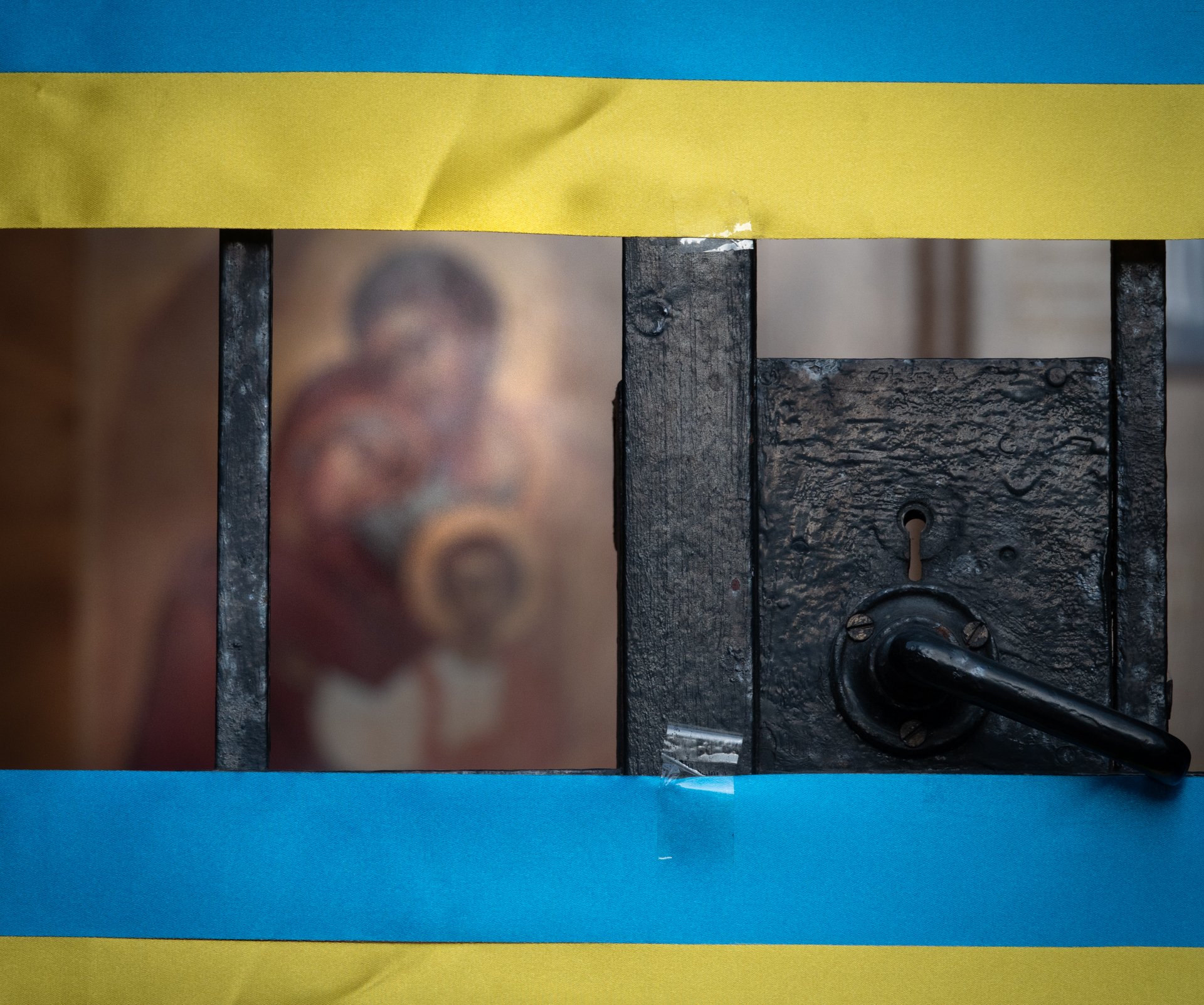 Blurred religious icon behind a wrought iron gate with a door handle, bordered by blue and yellow paper.