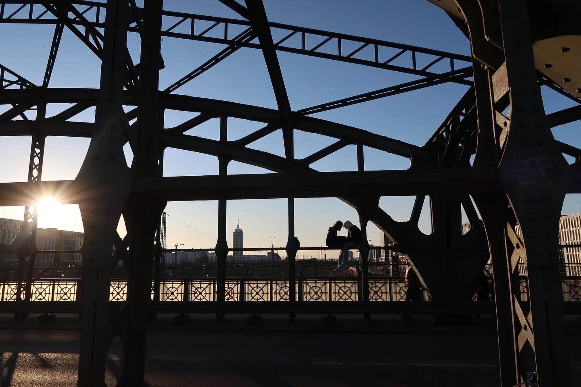 Silhouette of people sitting between iron beams of a bridge during sunset, with a city skyline in the background.