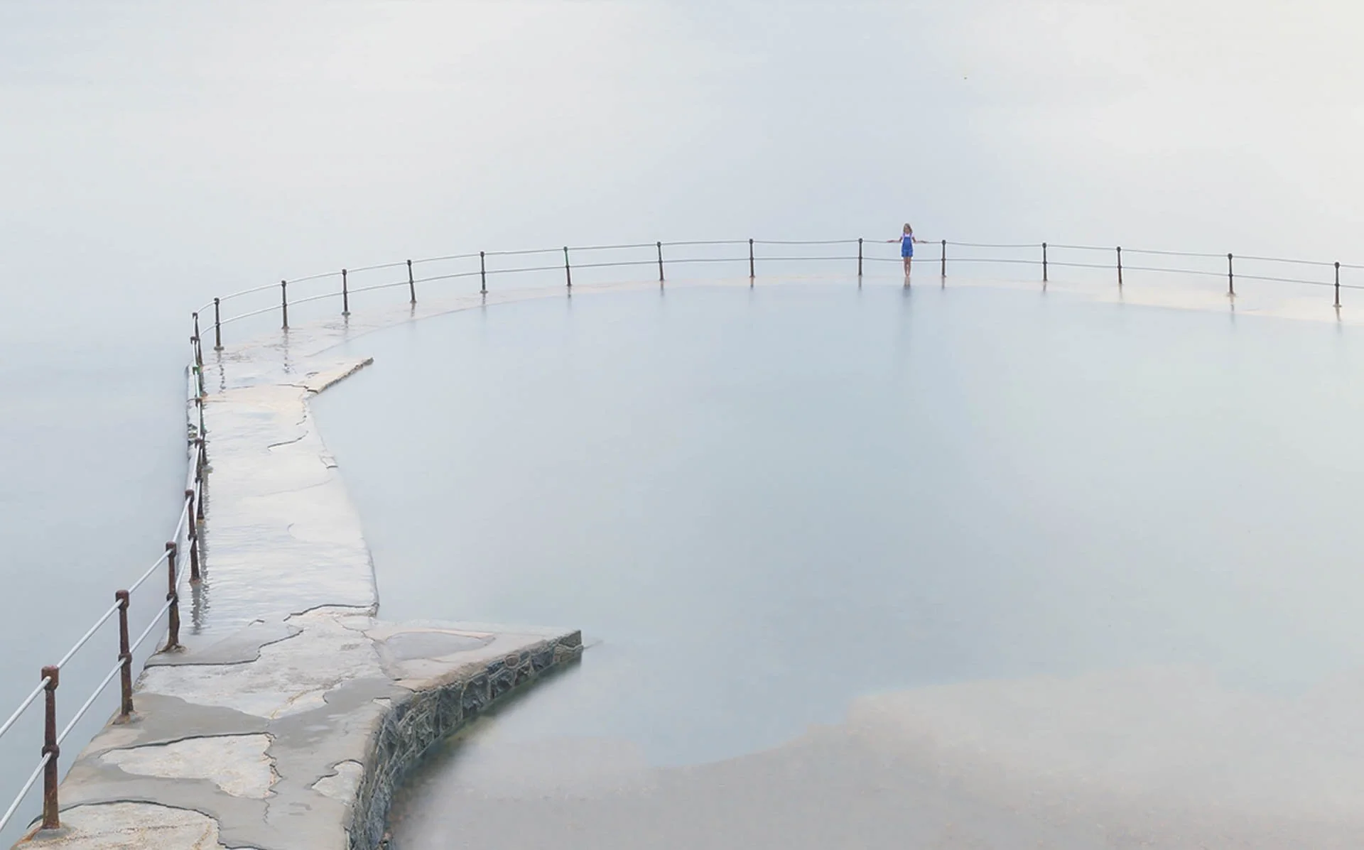 A solitary figure in blue stands on a curved, partially submerged walkway with metal railings, surrounded by calm, misty water.