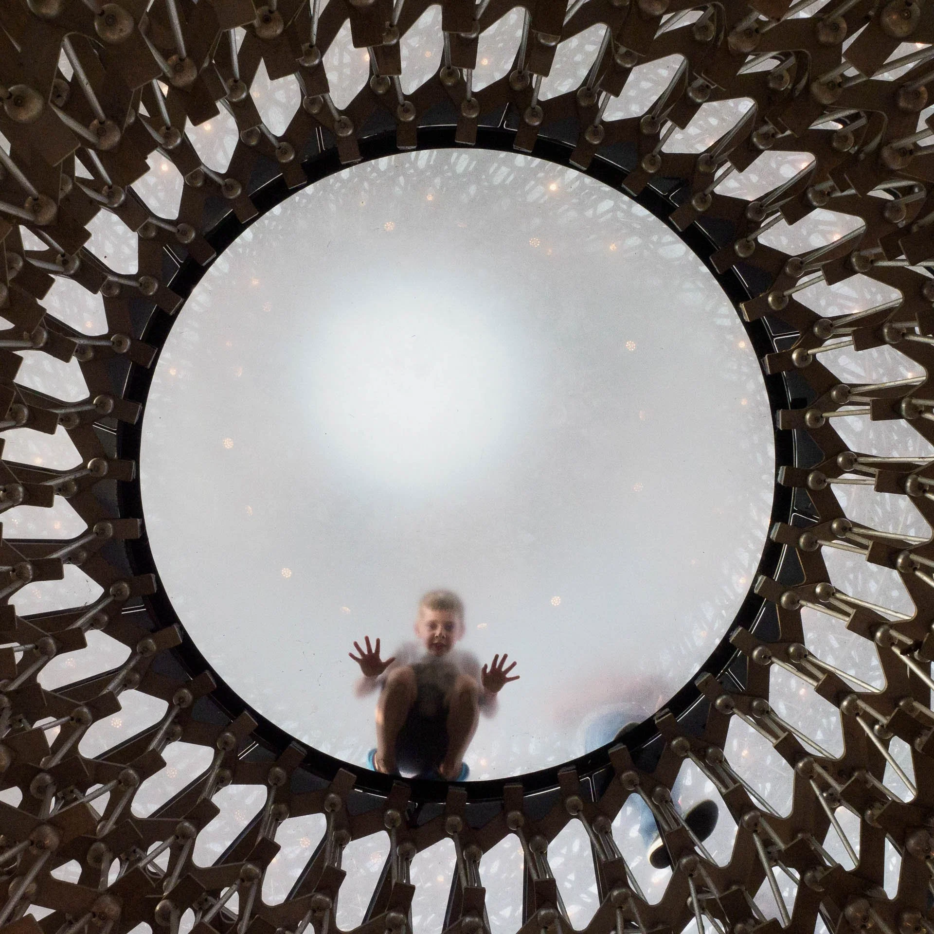 Child seen through a circular glass floor with intricate metal framework overhead.