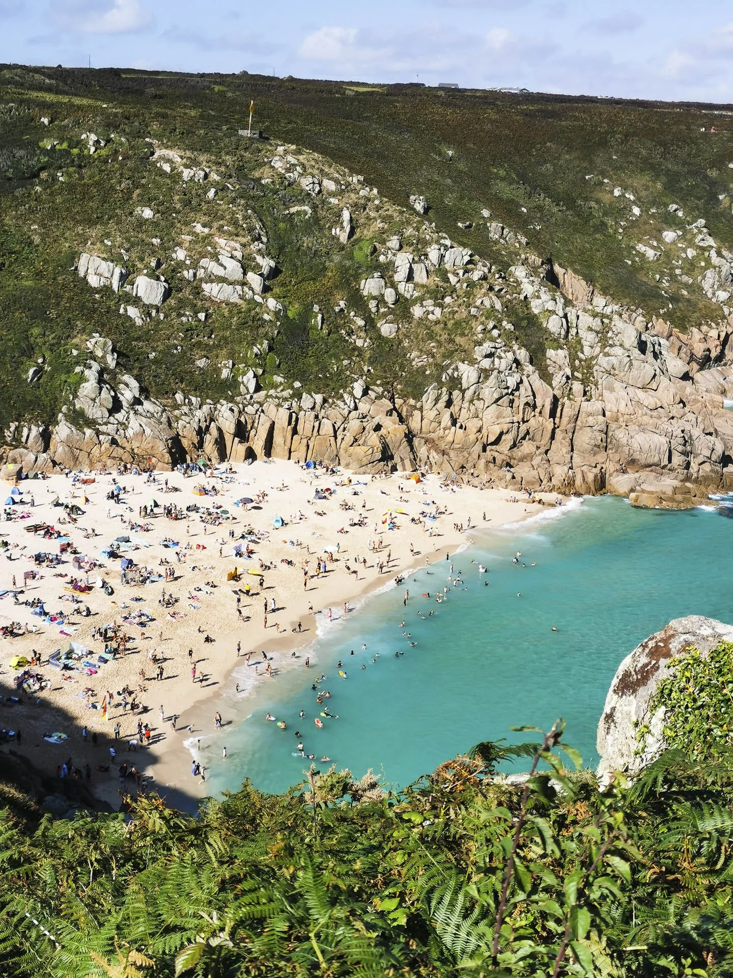 Aerial view of a crowded beach with turquoise water, surrounded by rocky cliffs and green vegetation.