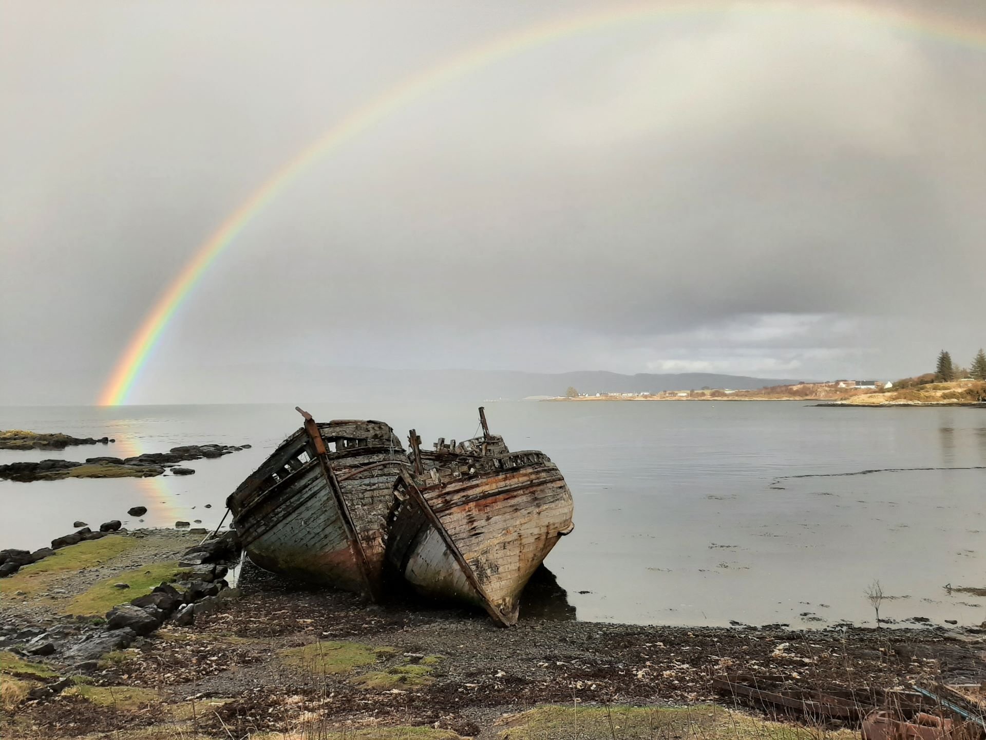 Two abandoned wooden boats on a rocky shore with a rainbow arching over calm water and a cloudy sky.