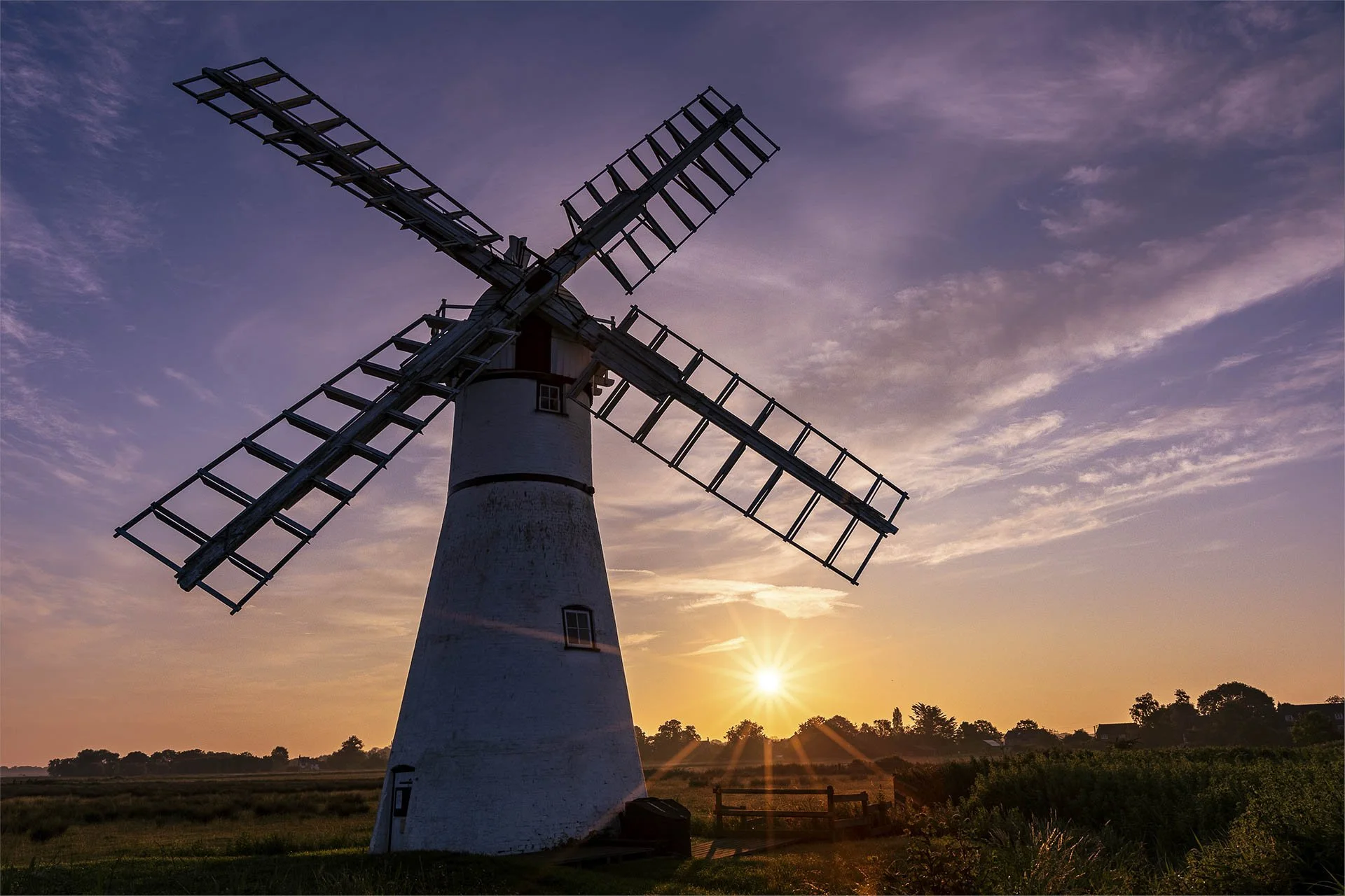 Silhouette of a traditional windmill at sunrise with sun rays, clear sky, and grassy landscape.