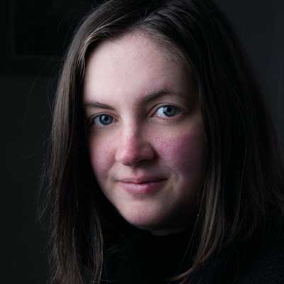 Close-up portrait of a woman with long brown hair looking at the camera against a dark background.