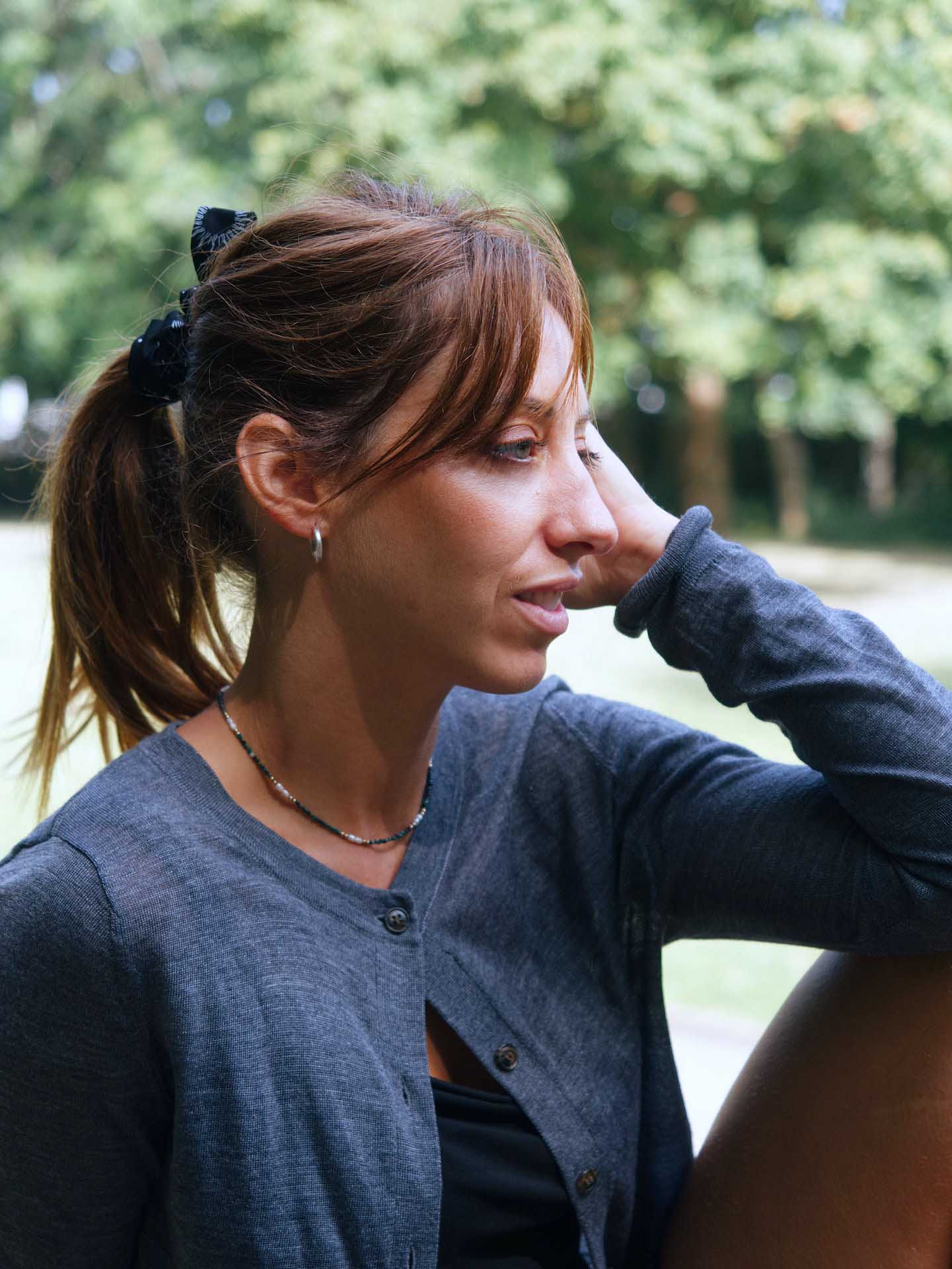 Woman with brown hair tied back, sitting outdoors in profile with her hand resting on her head, trees blurred in the background.