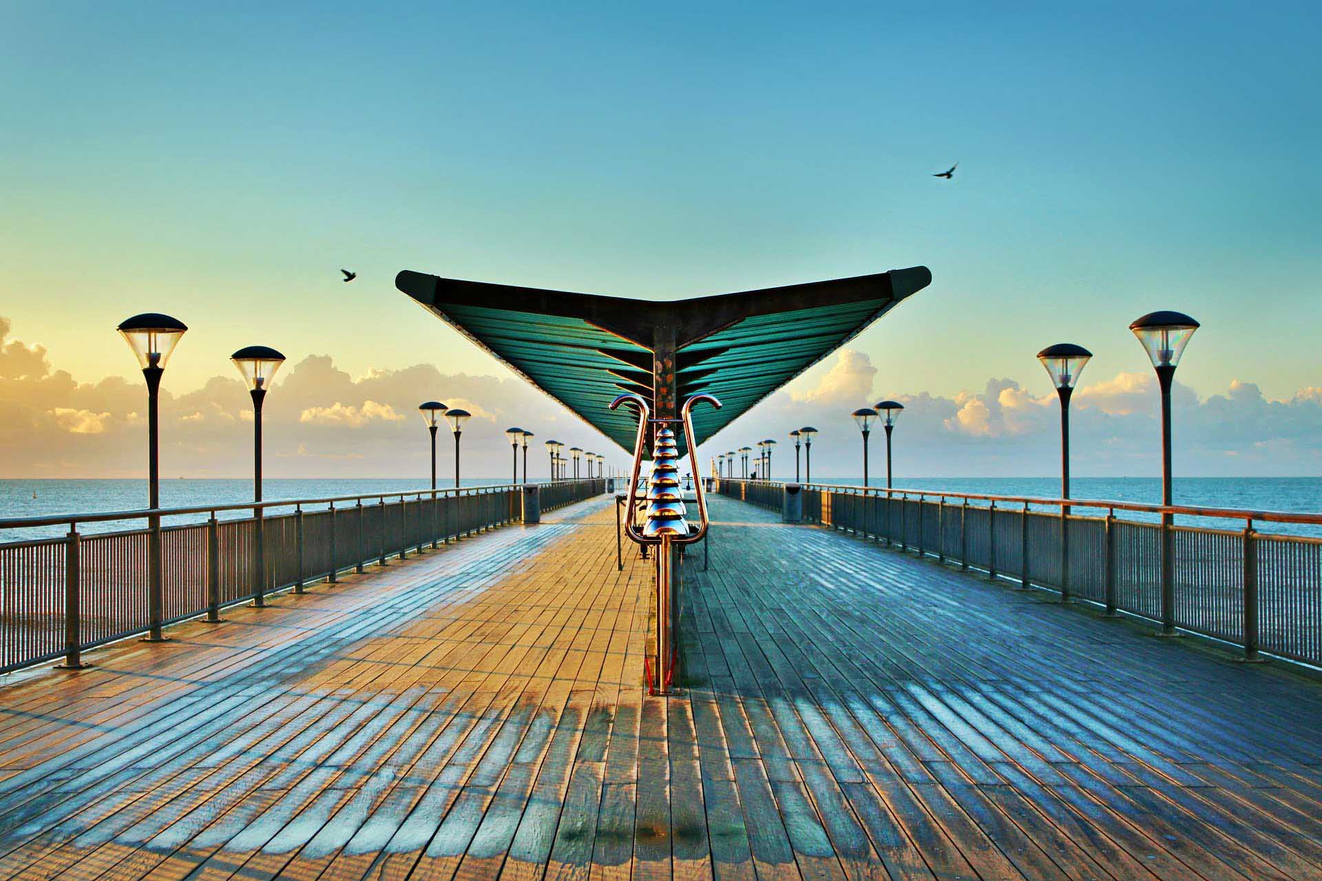 Symmetrical view down a wooden pier with modern lamps, centered sculptural canopy, calm sea on both sides, and soft clouds under a clear blue sky.