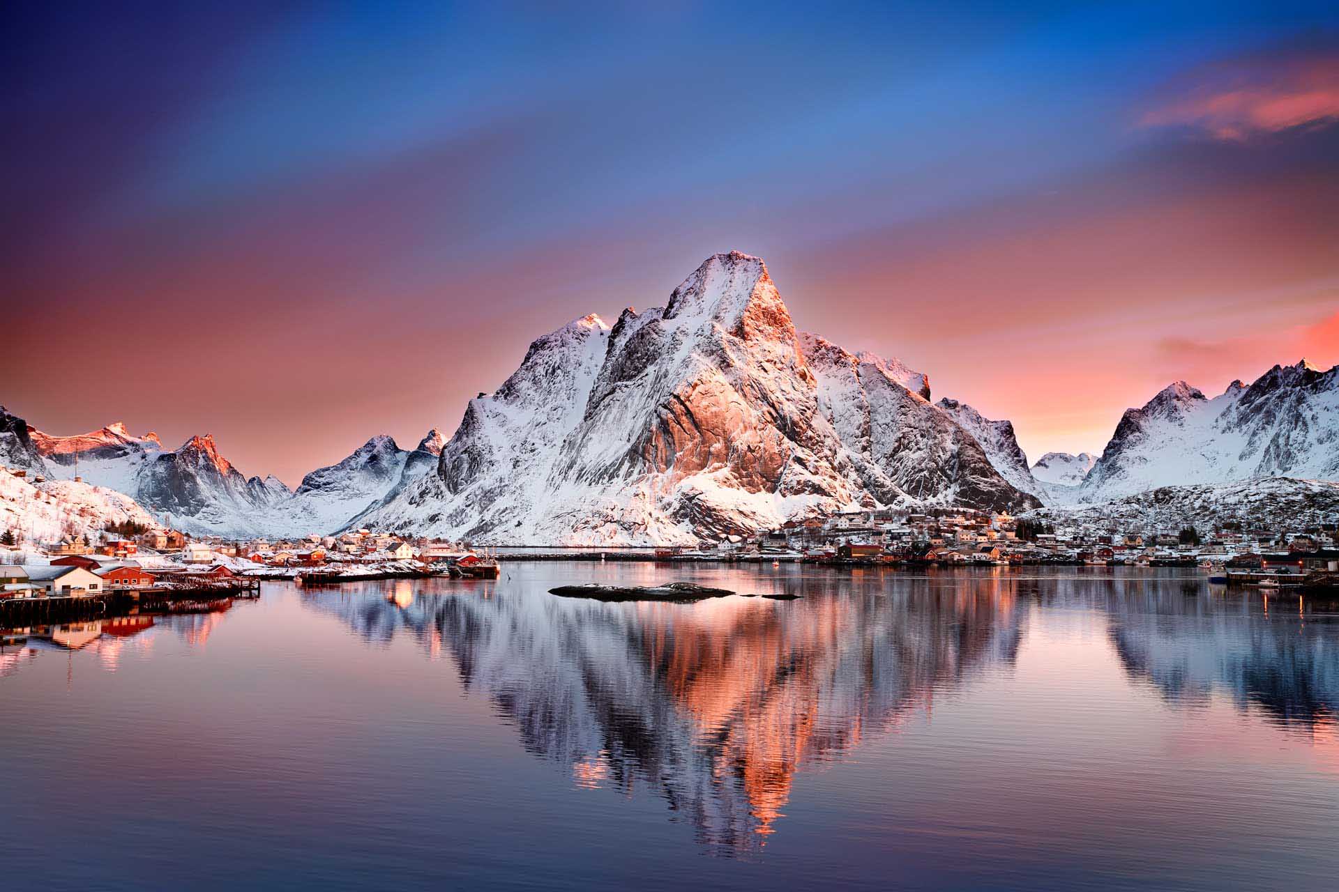 Snow-covered mountain rising above a coastal village at sunset, pastel sky reflected in still water.