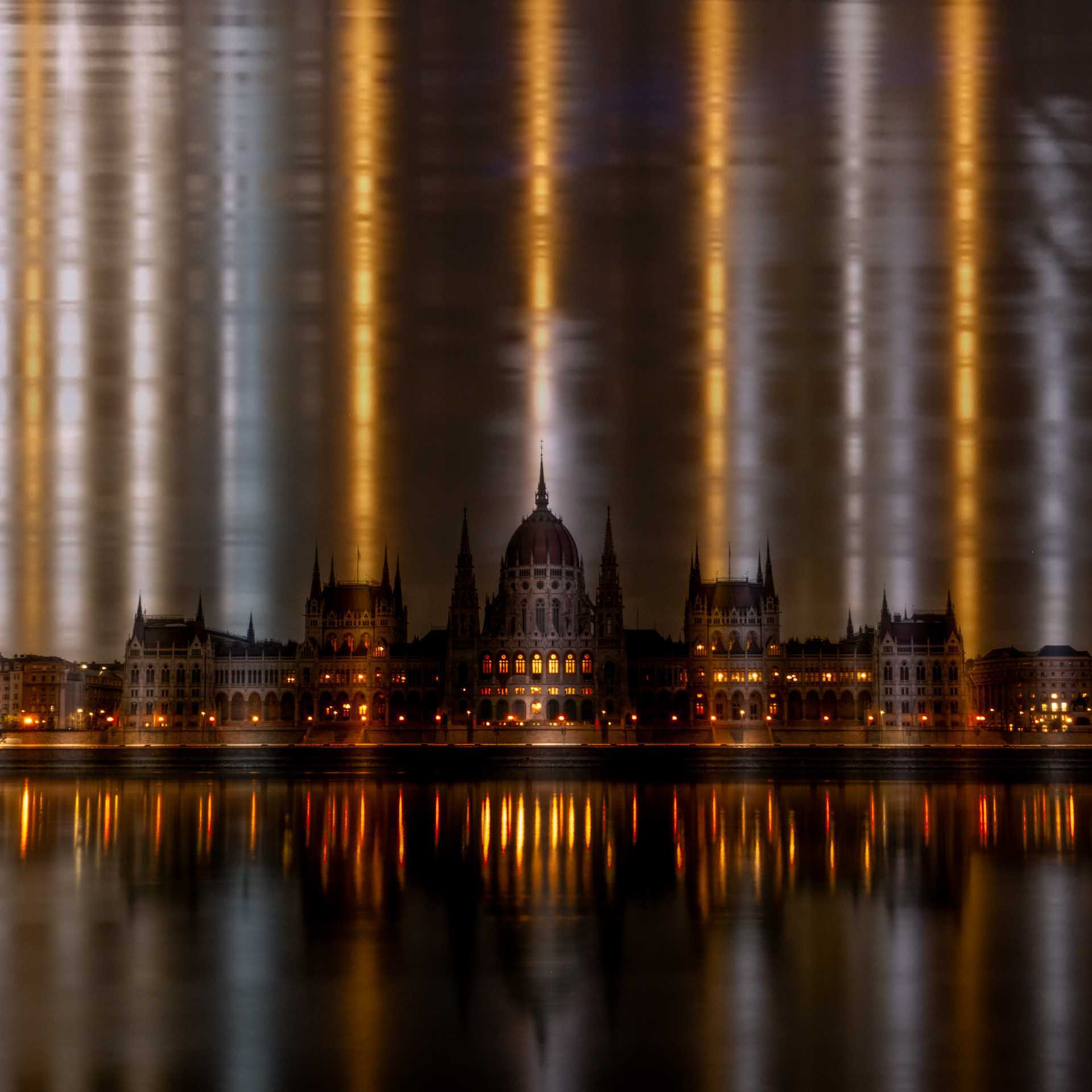 Illuminated grand parliament-style building at night with vertical light beams in the sky and reflections shimmering on the river.