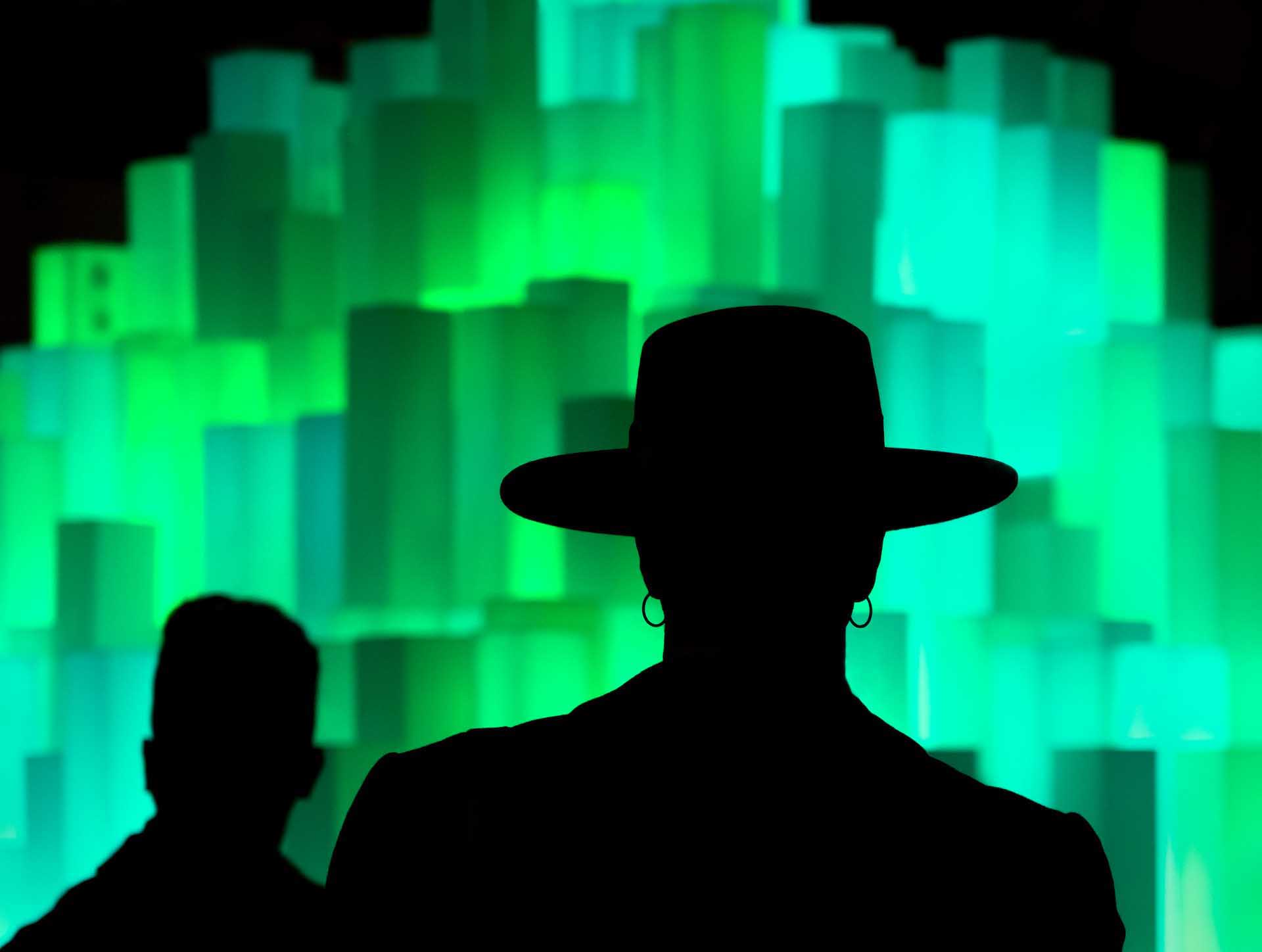 Silhouettes of two people facing a wall of glowing green rectangular light panels. One person is wearing a large hat.