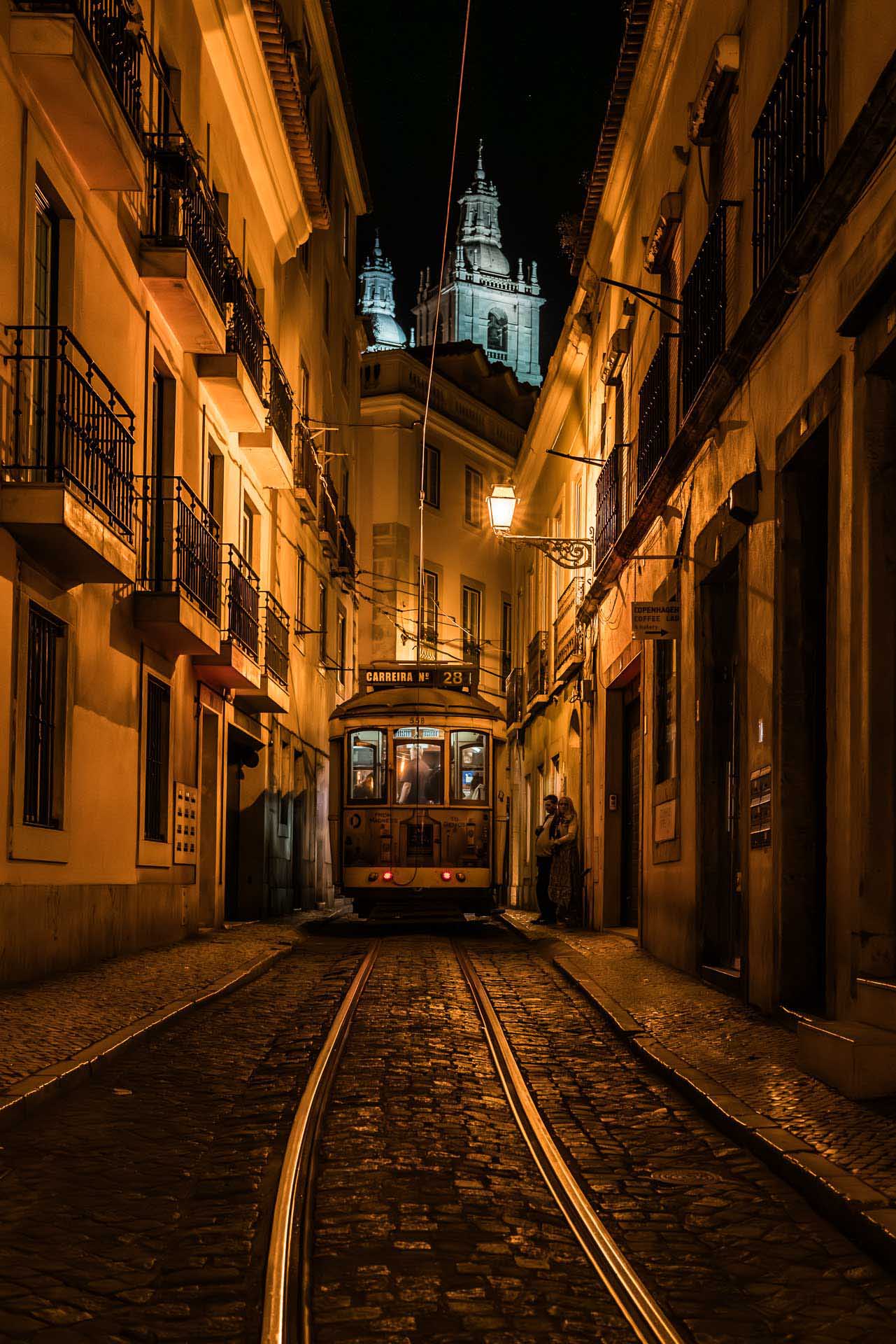 Yellow tram stopped on a narrow cobblestone street at night, framed by warm-lit buildings and overhead wires.