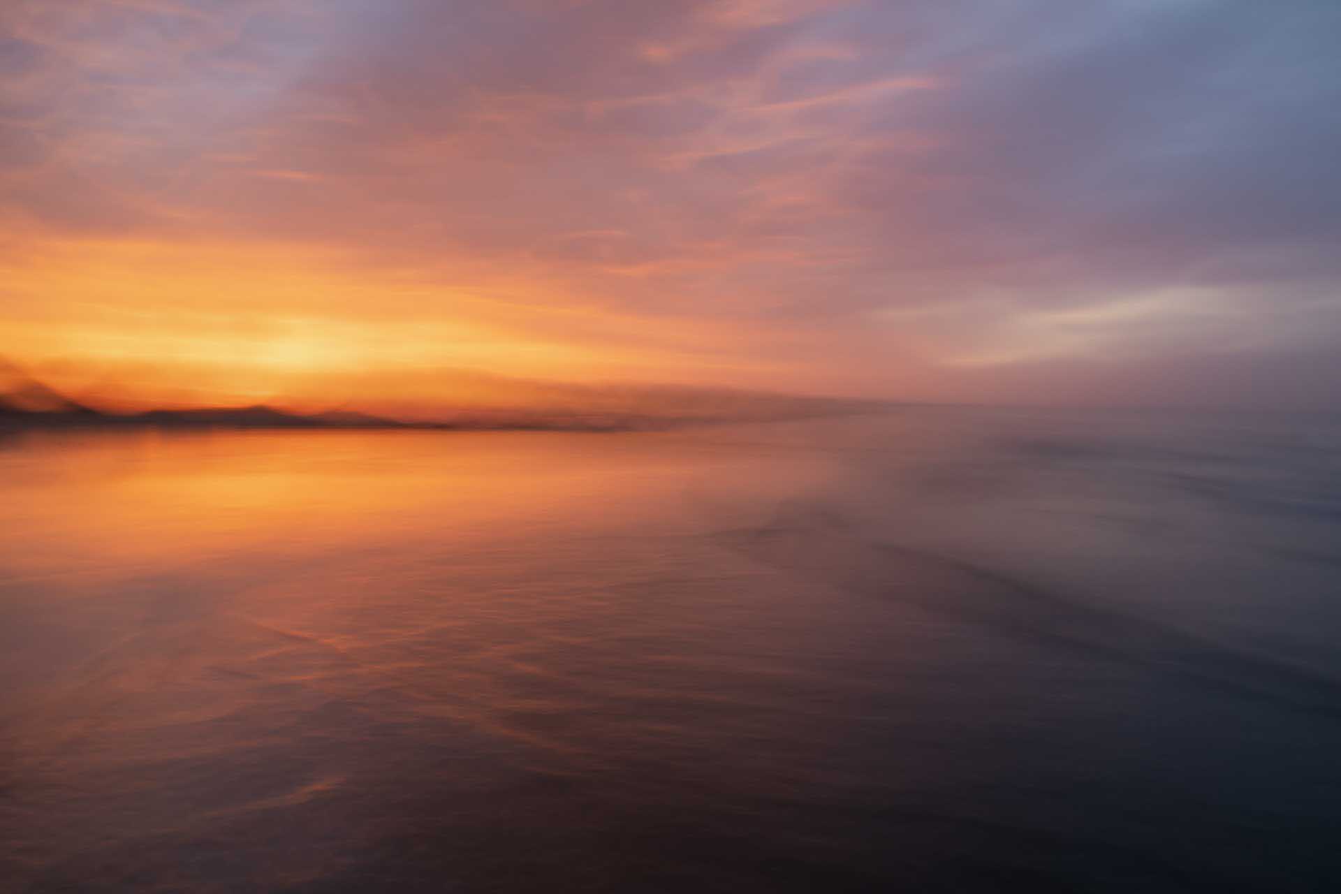 Soft, long-exposure sunset over calm water with blended orange and purple tones.