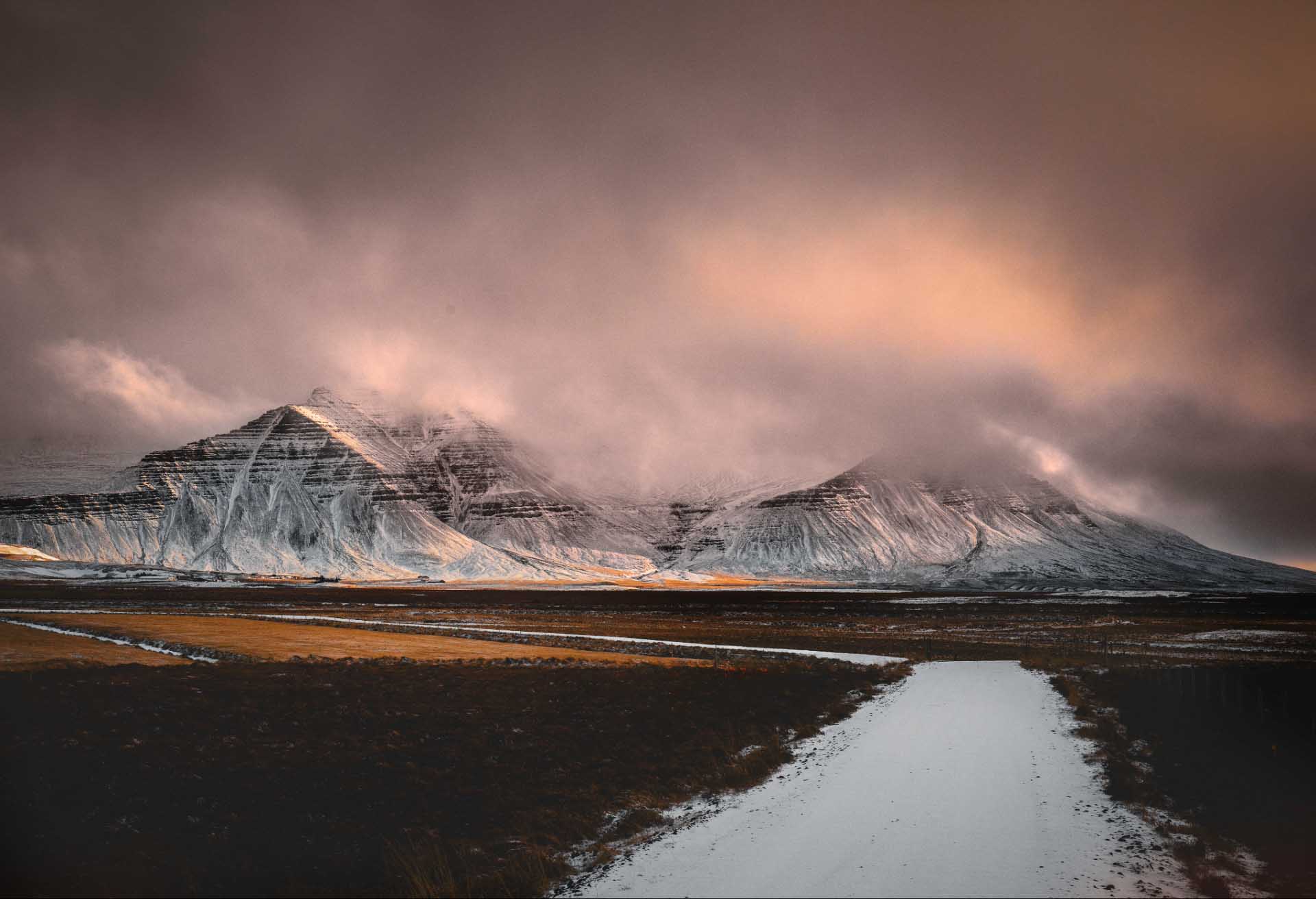 Snow-dusted mountains under low clouds with a narrow road cutting through a brown winter plain.