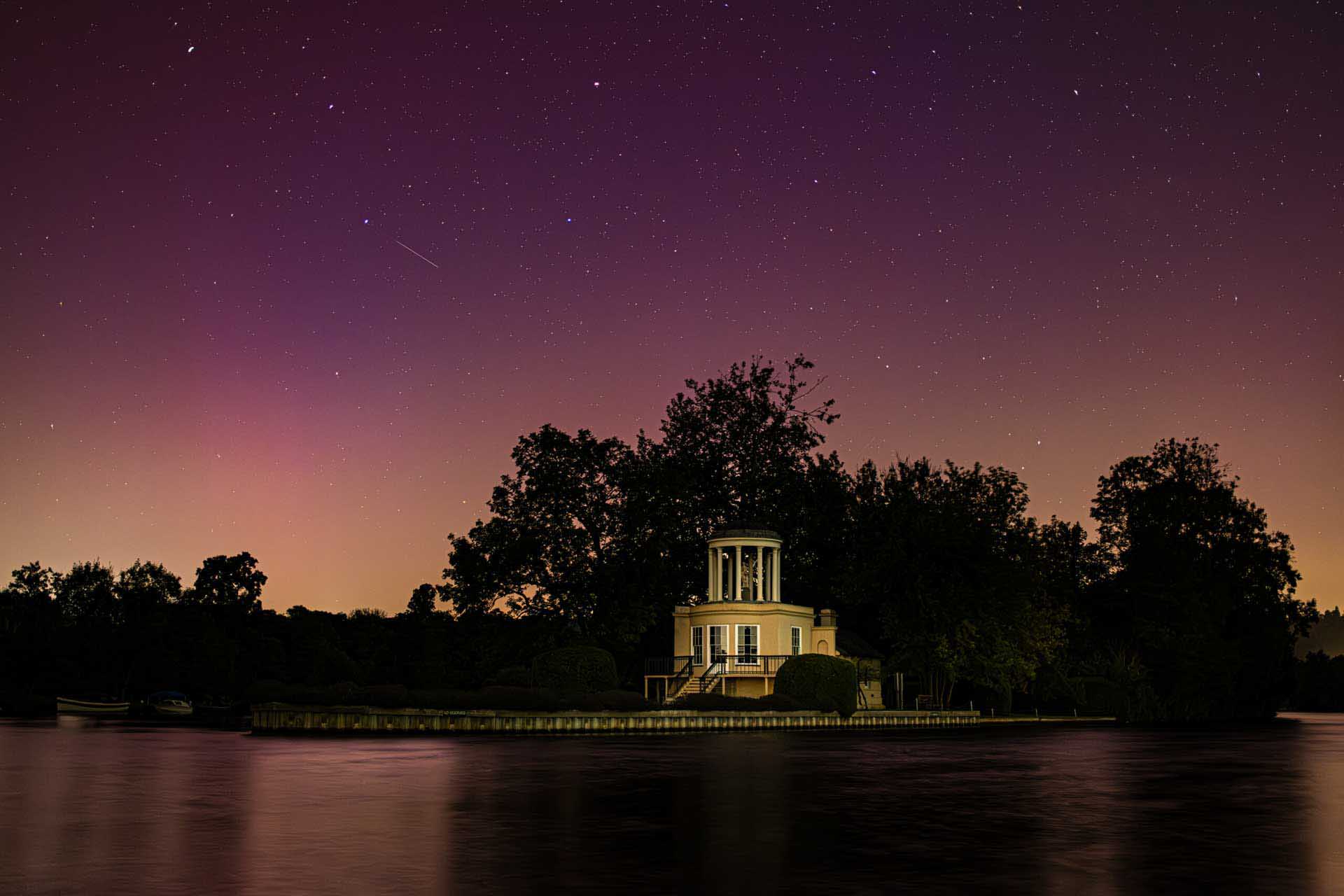 Small illuminated pavilion on an island, reflected in calm water beneath a star-filled purple sky.