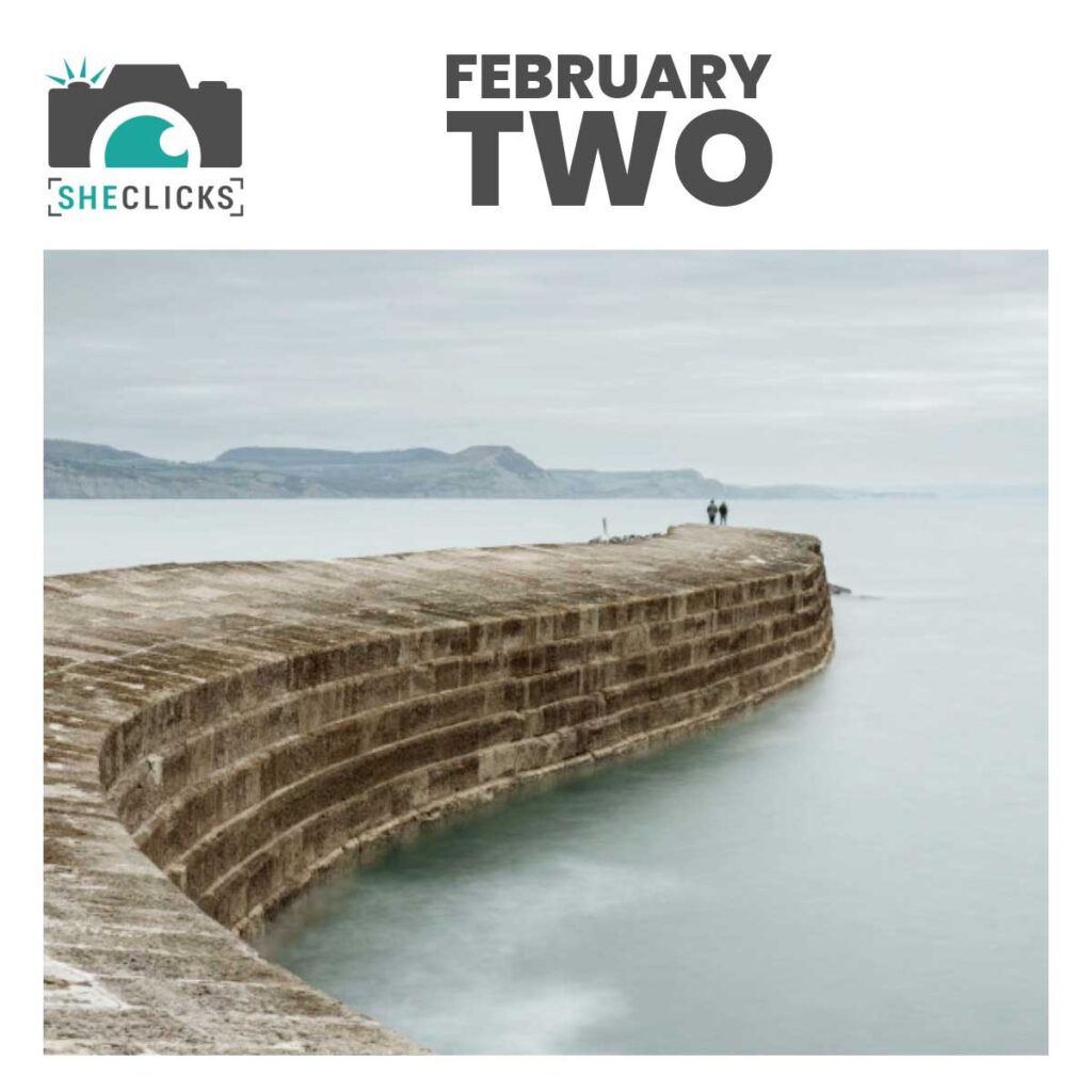 The curved stone pier of Lyme Regis Cobb extending into misty, blurred water with two small figures at the far end; overcast sky and distant hills in the background. Text at top reads “FEBRUARY TWO” with a SheClicks camera logo on the left.
