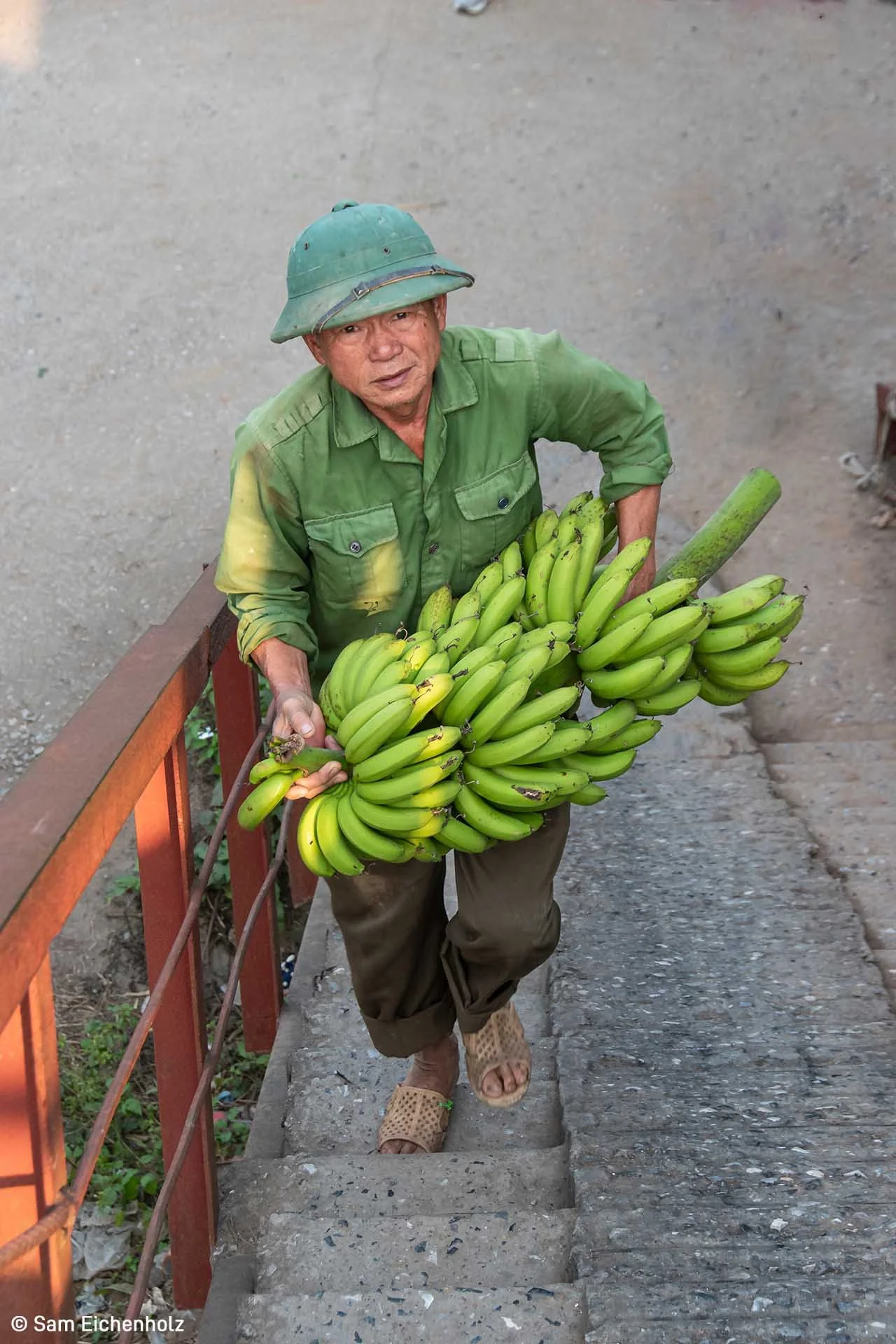  Image credit: Sam Eichenholz	Banana Haul	Under a bridge that spans the red river in Hanoi, Vietnam, residents have transformed public lands into bountiful gardens. As I descended the stairs to explore this hidden corner of the city, I came upon this man carrying his banana haul and was struck by the soulfulness of his eyes. 