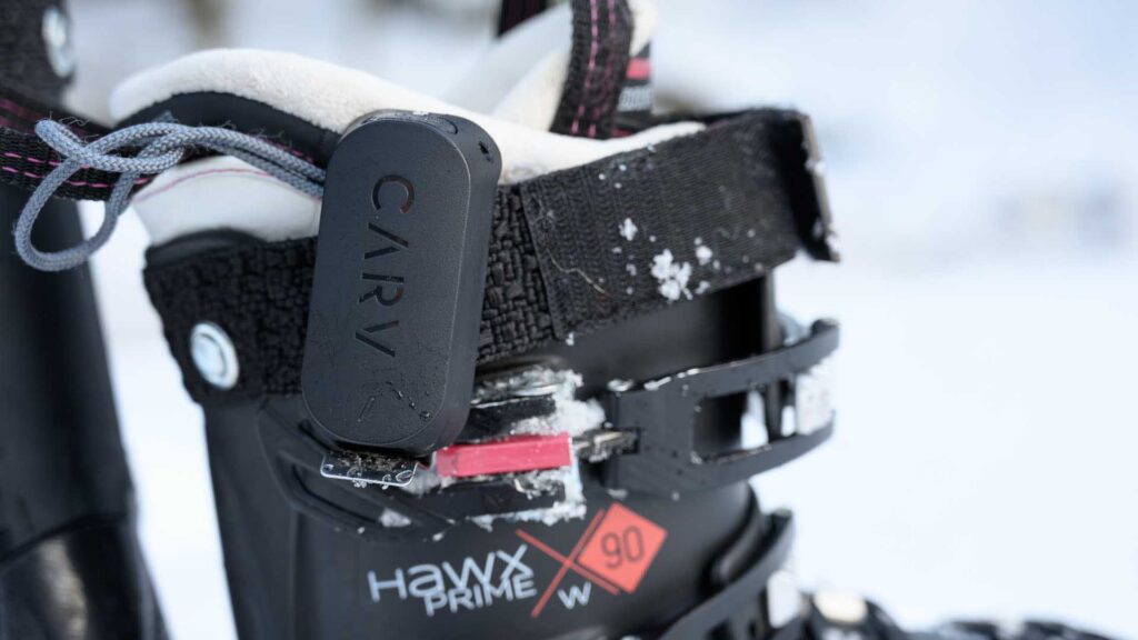 Close-up of the Carv 2 sensor clipped to a ski boot power strap, showing the embossed CARV logo with snow on the strap.