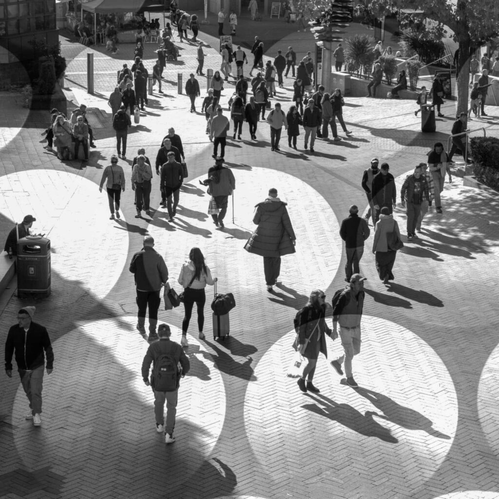 Black and white multiple exposure of people walking through a Birmingham city square, with overlapping circles of sunlight and long shadows across the paving.